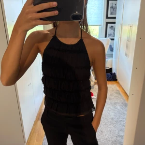 Svart halterneck topp med volanger - Säljer en svart halterneck topp med flera lager volanger och tunna knytband i ryggen. Toppen är ärmlös och har öppen rygg