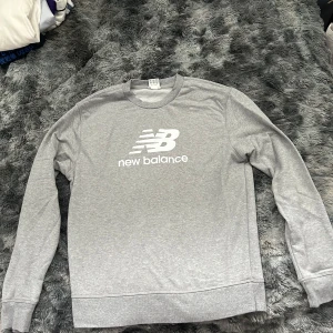 Grå sweatshirt från New Balance - Snygg grå sweatshirt från New Balance med klassisk logga i vitt framtill. Tröjan har rund halsringning och långa ärmar, perfekt för en chill och sportig stil. Tillverkad i mjuk bomullsmix som är skön att bära hela dagen.