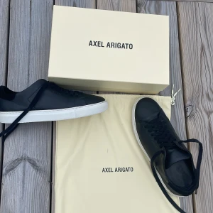 Svarta sneakers från Axel Arigato - Snygga svarta sneakers från Axel Arigato i skinn med vit platt sula och diskret guldfärgad logga på sidan. Klassisk låg modell med svarta snören och minimalistisk design. Skorna levereras med originalpåse och låda. Priset är inte hugget i sten och kan gå ner i sanbbaffär. Använda sparsamt i cirka en till två mån och kvitto finns köpta för cirka 2500. Killar  
