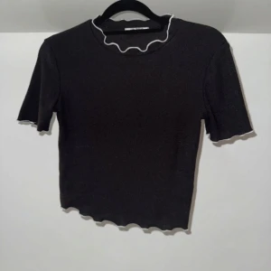 Svart ribbad topp med vita vågig kanter - Svart ribbad topp med kort ärm och croppad passform. Toppen har vågiga kanter vid hals, ärmslut och nederkant, med en vit kontrastkant som ger en snygg detalj. Perfekt för dig som gillar stilrena plagg med en twist.