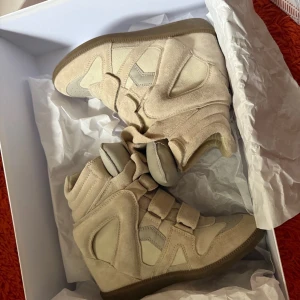 Beige Isabel Marant wedge sneakers - Säljer ett par ikoniska beige wedge sneakers från Isabel Marant. Skorna har coola paneler i mocka och skinn, kardborreband och en dold kilklack som ger extra höjd. Perfekta för dig som vill ha en edgy och trendig look. Kommer med originalkartong och dustbag.