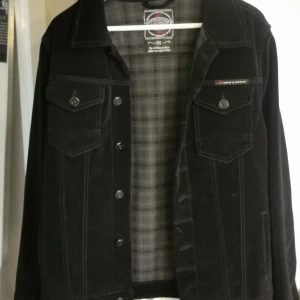 Svart manchesterjacka från Cipo & Baxx - Säljer en svart manchesterjacka från Cipo & Baxx med klassisk jeansjacke-look. Jackan har knappar framtill, två bröstfickor med lock och snygga sömdetaljer. Insidan är fodrad med rutigt tyg för extra stilpoäng. Perfekt för dig som vill ha en cool och tidlös jacka.