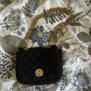 Svart axelväska med kedja och glitter - Snygg svart axelväska med glittrig yta och guldig kedjerem. Väskan har ett quiltat tyg och ett vridlås i guld framtill. Perfekt storlek för det viktigaste och ger en lyxig känsla till din outfit.