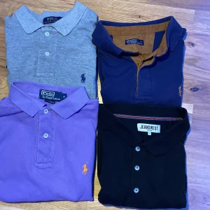 Fyra pikétröjor från Polo Ralph Lauren & Jeanswest - Säljer fyra klassiska pikétröjor: en grå, en marinblå med bruna detaljer, en lila och en svart. Tre från Polo Ralph Lauren med broderad logga och en från Jeanswest.gråa:300kr. Blåa 200kr. Lila 150kr. Svarta 50kr. För allt kan vi göra 500kr