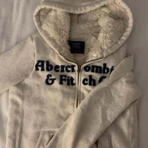  Abercrombie & Fitch hoodie - Mysig vit hoodie från Abercrombie & Fitch med fluffigt foder och stor huva. Tröjan har dragkedja framtill, broderad mörkblå logga och två fickor. Perfekt för kalla dagar när du vill vara både bekväm och snygg.