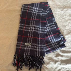 Rutig ullhalsduk från Burberrys - Klassisk rutig halsduk från Burberrys i mörka toner med inslag av rött, grått och svart. Halsduken är tillverkad i 100% ull och har fransar i ändarna. Perfekt accessoar för att lyfta din vinterstil.