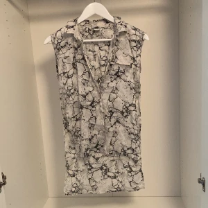 Ärmlös mönstrad kofta från Lindex - Snygg ärmlös kofta från Lindex i svartvitt abstrakt mönster. Kofta har krage, knappar framtill och en bröstficka. Lätt och luftig modell som passar perfekt för varma dagar. Liten rivning som lätt går att sy ihop.