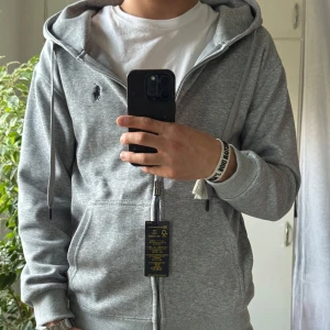 Grå hoodie från Polo Ralph Lauren - Snygg grå zip hoodie från Polo Ralph Lauren med dragkedja, huva och snörning. Tröjan har broderad logga på bröstet, kängurufickor och ribbade muddar. Slim fit-modell i mjuk bomullsblandning, perfekt för chill dagar.