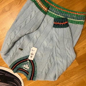 Ljusblå stickad tröja från Lacoste - Säljer en ljusblå stickad tröja från Lacoste med grov kabelstickning och färgglada ribbade muddar i grönt, blått, svart och orange. Tröjan har v-ringad hals och klassisk krokodil-logga på bröstet. Unisex och loose fit – perfekt för dig som gillar en chill stil.