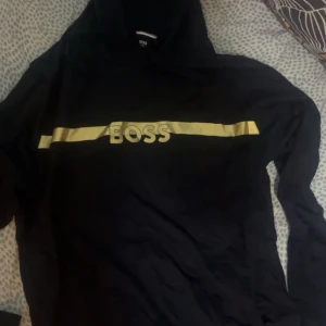 Svart hoodie från BOSS med gul detalj - Svart hoodie från BOSS med en bred gul rand och BOSS-logga i gult över bröstet. Tröjan har huva och långa ärmar, perfekt för en chill och stilren look. Klassisk passform och snygg kontrast mellan svart och gult.