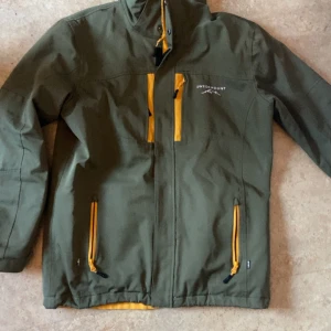 Grön vindjacka från Swedemount - Snyggt grön vindjacka från Swedemount med gula detaljer och dragkedjor. Jackan har hög krage, flera fickor med dragkedja och logga på bröstet. Perfekt för dig som gillar outdoor och vill ha en stilren jacka med coola kontrastfärger.