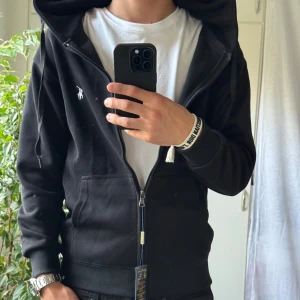 Svart hoodie från Polo Ralph Lauren - Svart zip hoodie från Polo Ralph Lauren med vit broderad logga på bröstet. Tröjan har dragkedja, huva med snörning och två stora fickor framtill. Tillverkad i mjuk bomull och har ribbade muddar vid ärmslut och midja. Perfekt för en chill och stilren look.
