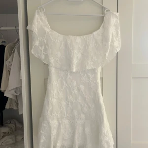 Vit blommig offshoulder klänning - Superfin vit kortklänning med blommigt spetsmönster och offshoulder-modell. Klänningen har volang upptill och är figurnära med en lätt utsvängd nederdel. Perfekt för sommarens alla tillfällen!