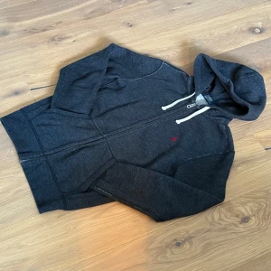 Mörkgrå hoodie från Polo Ralph Lauren - Mörkgrå hoodie från Polo Ralph Lauren med dragkedja, vit snörning i huvan och klassisk röd logga broderad på bröstet. Tröjan har långa ärmar, känguruficka och är tillverkad i mjuk bomullsblandning. Perfekt för chill dagar eller när du vill ha en clean streetstyle.