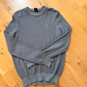 Grå tröja från Hugo Boss - Snygg grå tröja från Hugo Boss med diskret logga på bröstet. Tröjan har rund halsringning, ribbade muddar och är tillverkad i mjukt bomullsmaterial. Perfekt för dig som gillar stilren och enkel design. Size M men passar även S då den sitter väldigt tight