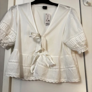 Vit blus med knyt från Gina Tricot - Supersöt vit blus från Gina Tricot med v-ringning och två knyt framtill. Blusen har korta puffärmar med spetsdetaljer och volangkant nertill. Perfekt för en romantisk look och tillverkad i mjuk bomull.