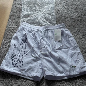 Vita badshorts från Lacoste med snörning - Snygga vita badshorts från Lacoste med stor svart krokodilprint på sidan och klassisk broderad logga framtill. Shortsen har elastisk midja med snörning och är tillverkade i lätt syntetmaterial, perfekta för sommaren och strandhäng.