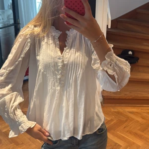 Vit romantisk blus med volangdetaljer - Supersöt vit blus med volangkant vid halsringningen och ärmslut. Blusen har knappar framtill, smockad överdel och långa, luftiga ärmar med rynkade detaljer. Perfekt för en bohemisk och avslappnad look. Tillverkad i ett lätt och skönt bomullstyg.