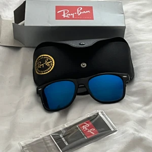 Ray-Ban Wayfarer solglasögon blå - Säljer ett par ikoniska Ray-Ban Wayfarer solglasögon med svart plastbåge och spegelblå linser. Modellen är RB2140, tillverkad i Italien. Kommer med originalfodral och putsduk. Perfekt för dig som vill ha en klassisk look med en modern twist.