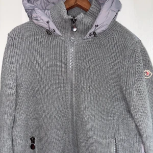 Grå cardigan jacka från Moncler - Säljer en grå cardigan jacka från Moncler med stickade ärmar och framsida, samt quiltad puffer-rygg och huva i ljusgrått. Jackan har dragkedja framtill, två sidofickor med dragkedja och Moncler-logga på ärmen. Perfekt för dig som gillar stilren och chill look. 2/M men passar S
