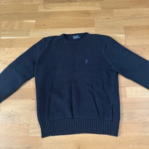 Svart stickad tröja Polo Ralph Lauren - Klassisk mörkblå stickad tröja från Polo Ralph Lauren med lila broderad logga på bröstet. Tröjan har rund halsringning, ribbade muddar och är långärmad. Perfekt för dig som gillar stilren och tidlös design.