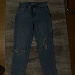 Blå högmidjade ripped jeans - Säljer ett par blå jeans med hög midja och slitna detaljer på båda benen. Jeansen har klassisk femficksdesign och raka ben. Perfekta för en avslappnad och trendig look.