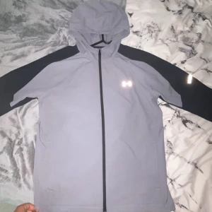 Grå vindjacka från Under Armour - Snygg grå vindjacka från Under Armour med svarta ärmar och hel dragkedja framtill. Jackan har huva och reflekterande logga på bröstet samt detaljer på ärmarna. Perfekt för blåsiga dagar och sportiga outfits