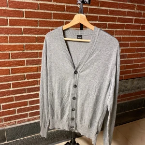 Grå kofta från Hugo Boss med knappar - Snygg grå kofta från Hugo Boss med v-ringning och svarta knappar framtill. Tröjan har ribbade muddar och är långärmad, perfekt för lager-på-lager. Tillverkad i mjuk bomullsmix som känns skön mot huden.