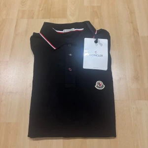 Svart pikétröja från Moncler - Svart pikétröja från Moncler med klassisk krage och knäppning framtill. Tröjan har Moncler-logga på bröstet och röda samt vita ränder på krage och ärmslut. Tillverkad i bomull för en skön och stilren känsla.