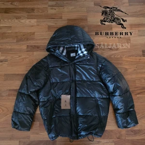 Burberry Pufferjacka - Svart dunjacka från Burberry med klassisk Säljer en elegant och mångsidig Burberry-jacka i svart, i nyskick. Dunfodrad för värme och komfort med det klassiska rutmönstret i huvan som ger en exklusiv touch. Praktiska dragkedjefickor, avtagbar huva och diskret logopatch på ärmen. Sitter som (M/L). Jackan är i princip som ny och utan defekter. En lyxig kombination av stil och funktion – perfekt för både vardag och tillfällen då du vill klä upp dig.  📦Snabb leverans - skickas samma dag📦 