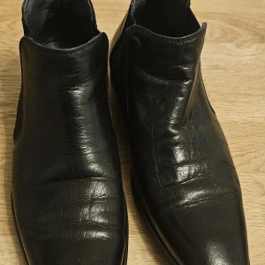 Svarta chelsea boots i skinn, stl 40 - Snygga svarta chelsea boots i äkta skinn med spetsig tå och elastiska paneler på sidorna. Klassisk och stilren modell som passar perfekt till både jeans och kostym. Platt sula och låg klack för en clean look.