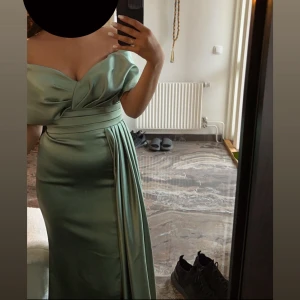 Grön långklänning i satin med slits - Elegant grön långklänning i glansigt satinmaterial med offshoulder-skärning och markerad midja. Klänningen har en snygg slits framtill och draperade detaljer upptill som ger en lyxig känsla. Perfekt för dig som vill sticka ut med en sofistikerad look. 