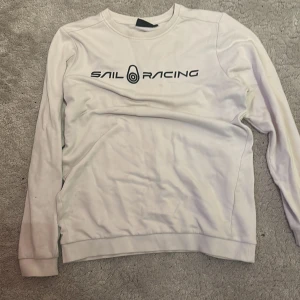 Vit Sail Racing sweatshirt - Vit långärmad sweatshirt från Sail Racing med svart logga tryckt på bröstet. Klassisk rund halsringning och ribbade muddar vid ärmslut och nederkant. Perfekt för en clean och sportig stil.