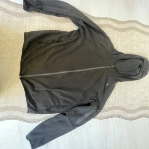 Svart hoodie från Björn Borg med dragkedja - Svart hoodie från Björn Borg med hel dragkedja framtill och huva. Tröjan har en diskret logga på bröstet och en praktisk ficka med dragkedja på ena ärmen. Tillverkad i mjuk bomull, perfekt för chill eller träning.