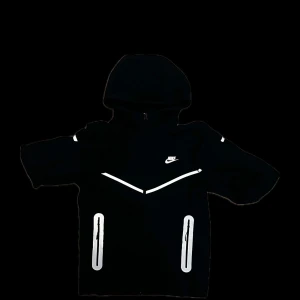Svart Nike vindjacka med reflexdetaljer - Svart vindjacka från Nike med huva och hel dragkedja. Jackan har vita reflexdetaljer på bröstet, ärmarna, fickorna och ryggen. Snygg sportig look med två fickor framtill och Nike-logga på bröstet. Perfekt för dig som gillar streetwear och vill synas i mörkret. Storlek S men passar som M.