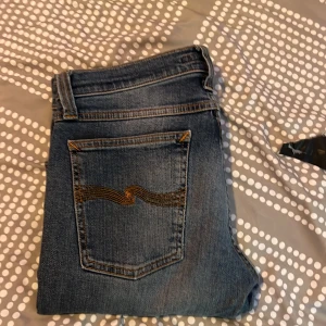 Blå jeans från Nudie Jeans med rå kant - Säljer ett par klassiska blå jeans från Nudie Jeans med snyggt slitna detaljer och rå, fransig kant nertill. Jeansen har fem fickor, orange sömmar och en rak passform. Perfekta för dig som gillar en avslappnad och stilren look.