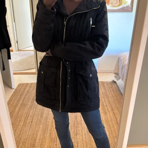 Svart parkajacka med bälte och huva - Svart värm parkajacka med huva, dragkedja och snyggt bälte i midjan. Jackan har flera fickor med knappar och dragkedjor, vilket ger en cool och praktisk look. Perfekt för dig som gillar streetstyle och vill ha en jacka som funkar hela säsongen.