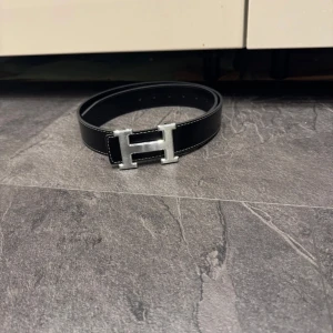 Svart  skärp med H-spänne - Snyggt svart bälte med ikoniskt H-spänne i silverfärgad metall. Bältet är gjort i äkta skinn med vit kontrastsöm längs kanterna och har en stilren, klassisk design som lyfter vilken outfit som helst.