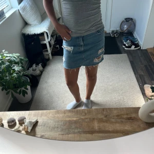 Blå jeanskjol med slitningar - Säljer en kort blå jeanskjol med råa kanter och slitna detaljer framtill. Kjolen har klassisk femficksdesign och dragkedja med knapp. Perfekt för dig som gillar en avslappnad och trendig stil.