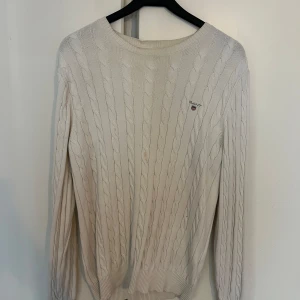 Vit kabelstickad tröja från GANT - Vit kabelstickad tröja från GANT med broderad logga på bröstet. Klassisk rund halsringning och ribbade muddar. Perfekt för dig som gillar preppy stil och vill ha en tidlös tröja i garderoben.
