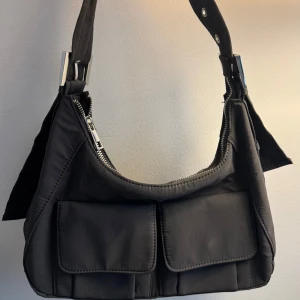 Grå axelväska med fickor och dragkedja - Grå axelväska i syntetmaterial med två stora fickor framtill och silvriga metalldetaljer. Väskan har justerbart axelband med spänne och dragkedja upptill. 