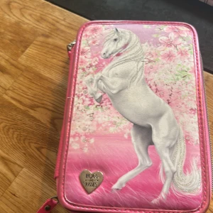 Rosa plånbok med hästmotiv Hugs & Kisses - Supergullig rosa plånbok med glittrande hästmotiv och blommor på framsidan. Insidan är dekorerad med små hästar och har flera fack och resårband för att hålla ordning på småsaker. Perfekt för dig som älskar hästar och vill ha något unikt!