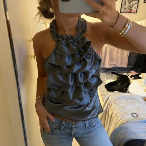 Satin volang topp - Supersnygg grå halterneck topp med flera volanger framtill. Toppen har en glansig satinliknande finish och knyts i nacken. Perfekt för dig som vill sticka ut med en cool och trendig look.