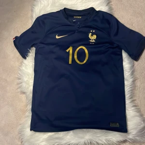 Frankrike Mbappe #10 VM 2022 tröja  - Snygg marinblå Frankrike fotbollströja från Nike med guldiga detaljer, nummer 10 och Mbappé på ryggen. Tröjan har korta ärmar, broderat landslagsemblem med tupp och två stjärnor på bröstet. Tillverkad i lätt och ventilerande polyester.