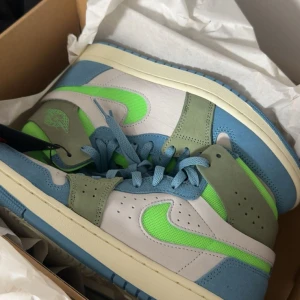 Nike Air Jordan 1 blå/grön sneakers - Säljer ett par Nike Air Jordan 1 sneakers med fräscha blå och gröna detaljer. Skorna har en vit bas, ljusblå mockadetaljer och neongröna swoosh-loggor. Klassisk hög modell med snörning och rund tå. Perfekta för dig som vill sticka ut med färg på fötterna.