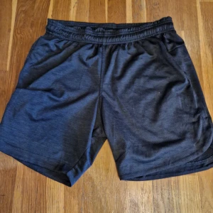 Svarta träningsshorts med resår - Säljer ett par svarta träningsshorts med elastisk midja. De har en loose passform och är tillverkade i ett lätt syntetmaterial som andas. Perfekta för gymmet eller löprundan. Snygg enkel design utan mönster eller extra detaljer.
