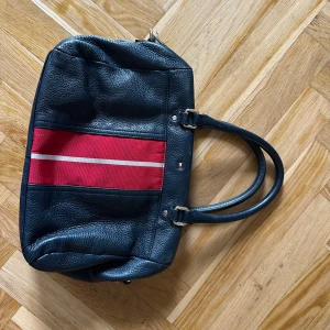 Tommy Hilfiger marinblå handväska - Snygg marinblå handväska från Tommy Hilfiger i skinn med röda och vita ränder framtill. Väskan har dubbla handtag och klassisk TH-logga på framsidan. Perfekt storlek för att få plats med allt du behöver och ger en sportig men stilren vibe.