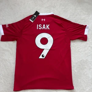Liverpool Isak #9 Adidas matchtröja - Liverpool FC fotbollströja från Adidas med Isak och nummer 9 på ryggen. Röd med vita detaljer, broderad Adidas-logga, Premier League-märke i guld på ärmen och Standard Chartered-tryck på bröstet. Tillverkad i ventilerande polyester för maximal komfort.