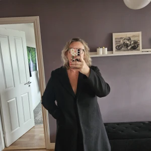 Svart kappa med klassisk krage - Säljer en stilren svart kappa med klassisk krage och rak passform. Jackan har långa ärmar och sidfickor, perfekt för dig som gillar en enkel och tidlös look. Passar dig som vill ha en snygg och clean siluett. 3 Tryck knappar framtill. 