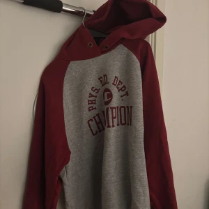 Grå och vinröd hoodie från Champion - Säljer en snygg hoodie från Champion i grått med vinröda ärmar och huva. Tröjan har tryck på bröstet, klassisk känguruficka och är perfekt för chill dagar. Mjuk bomullsblandning och relaxed fit. Passar dig som gillar sportig och avslappnad stil.